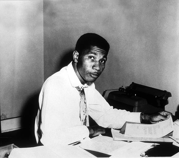 A black and white photo of Medgar Evers sitting at his desk. Evers is holding a sheet of paper. A typewriter can be seen behind him. Several papers are scattered across the desktop.