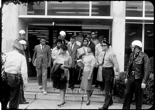 A black and white photograph of the Tougaloo Nine being escorted out of the Jackson Public Library. Two women lead the group as the others descend the two steps to the entrance. Three officers are on the left, two are in the rear, and two are on the right of the demonstrators.