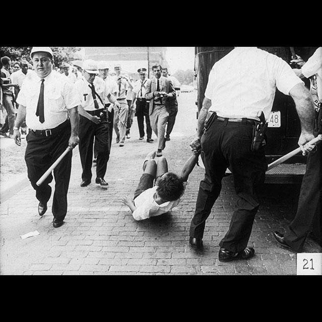 A black and white photograph of a woman being dragged by a police officer down a sidewalk to a police bus